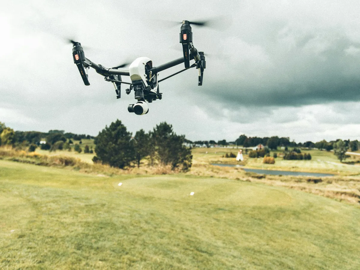 Duży biały dron z kamerą wisi nisko nad trawiastą łąką, w tle kępy drzew i pochmurne niebo