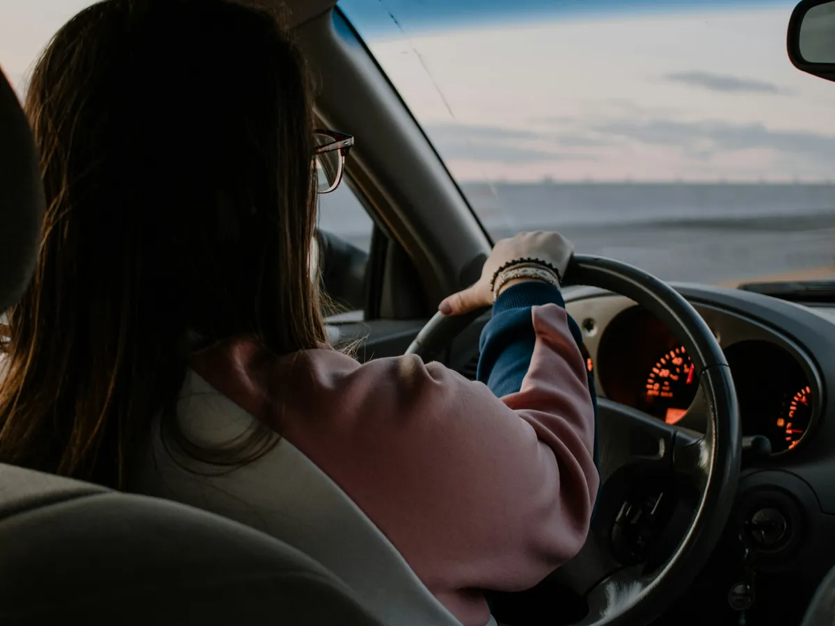 Kobieta w okularach i różowej bluzie prowadzi samochód, widok zza fotela kierowcy. Prawą ręką trzyma kierownicę, za szybą szare niebo i otwarta przestrzeń.
